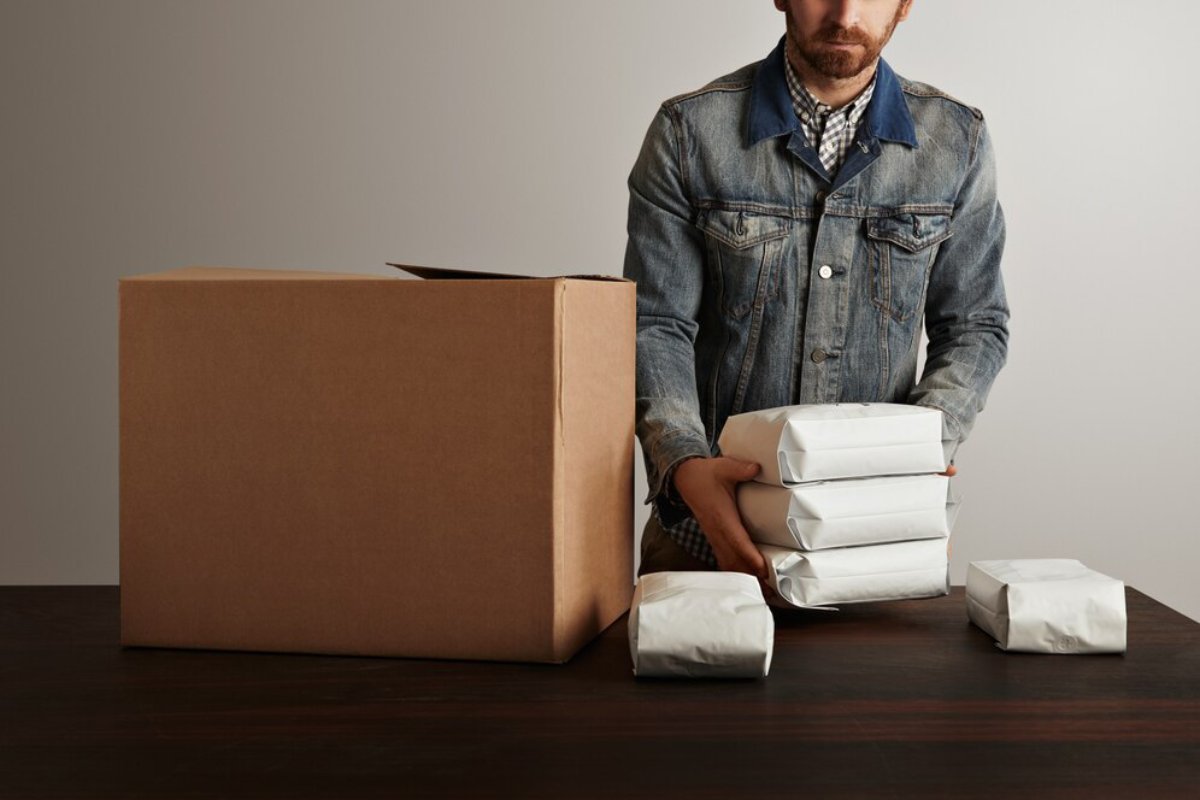 Man in a denim jacket packing and organizing several white parcels next to a large cardboard box on a table.