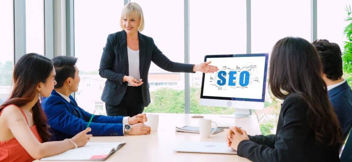 Businesswoman presenting SEO concepts on a computer screen to a group of attentive coworkers in an office setting.