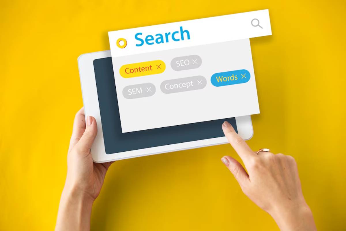 Hands holding a tablet displaying a digital search interface with keywords related to online content strategy against a yellow background.