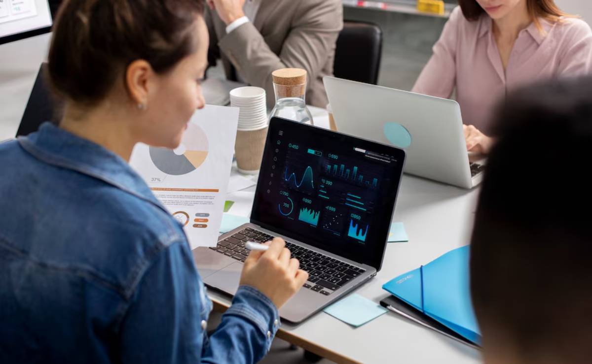 Business professionals engaged in a discussion, with one analyzing financial data on a laptop screen.