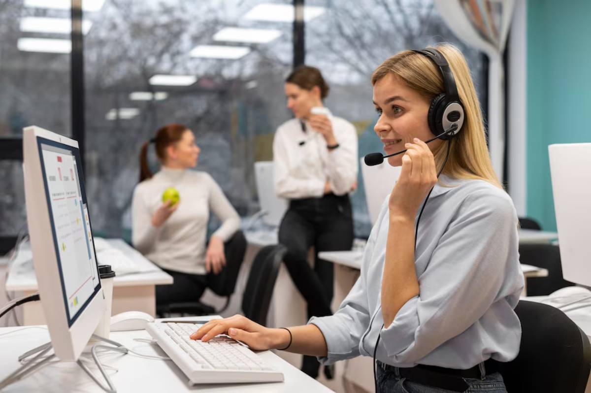 A customer service representative with a headset working at a computer in a busy office environment.