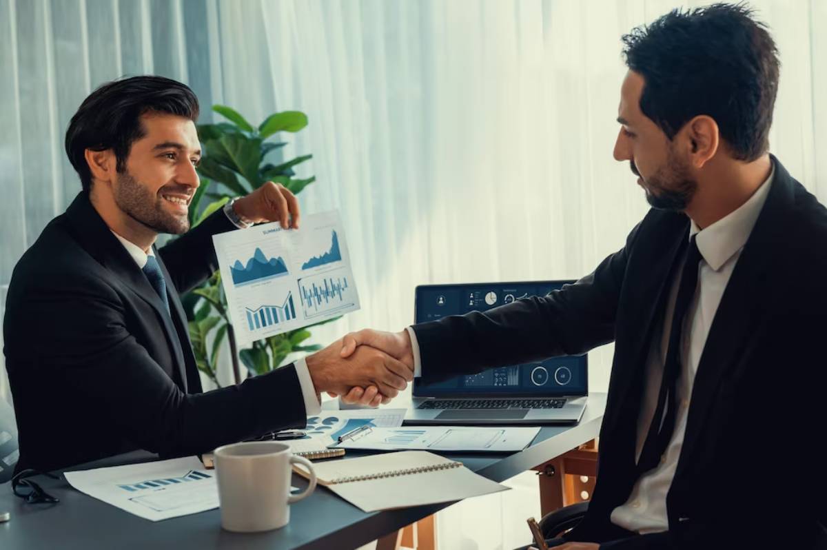 Two businessmen shaking hands in an office while discussing financial charts.