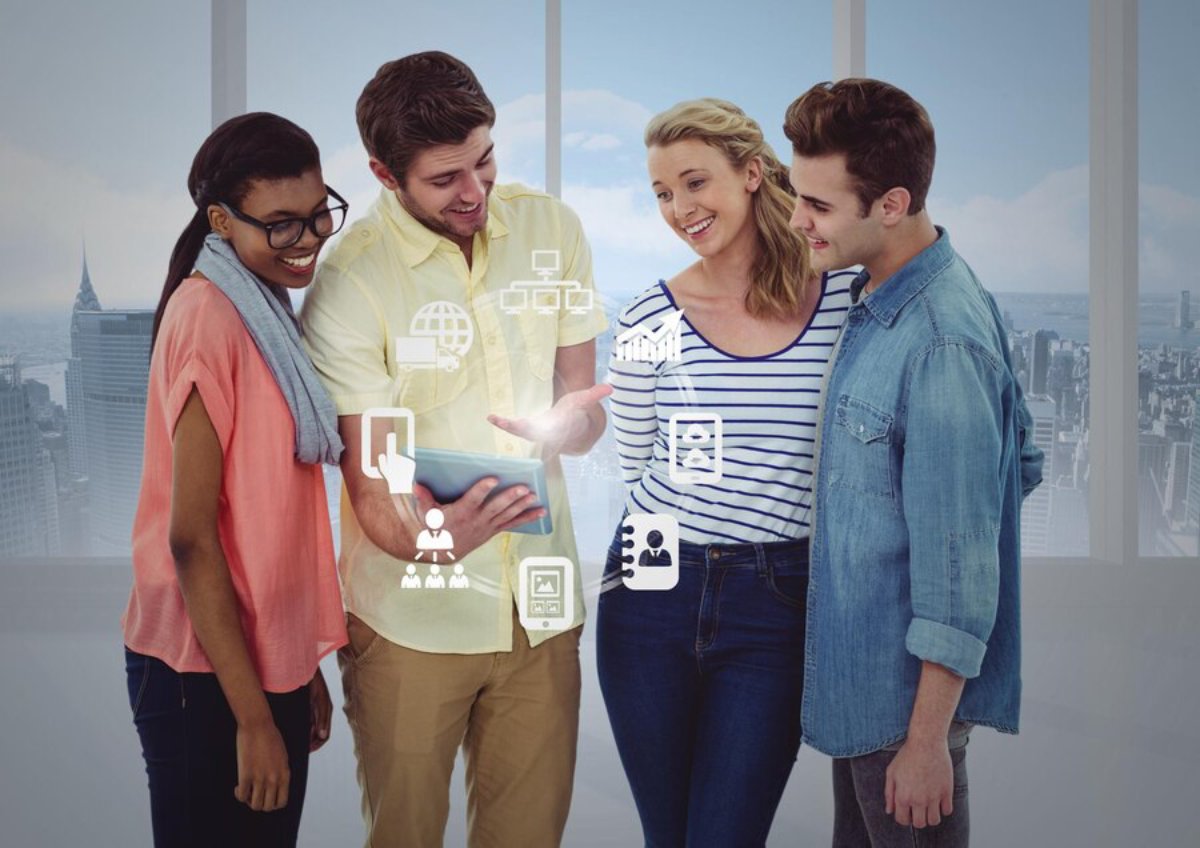 Four young adults examine a tablet together, displaying digital icons, in a modern office overlooking a cityscape.
