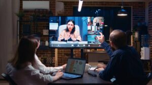 Two people participating in a video conference with multiple remote attendees displayed on a large screen in a modern office.