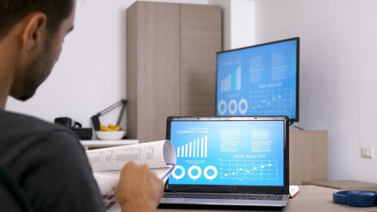 Man reviews financial documents while multiple screens display business data analysis graphs in a modern office setup.