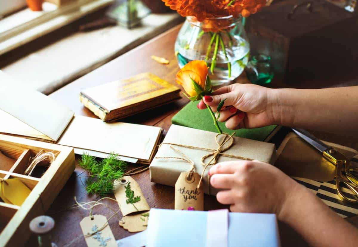 Person crafting with books, gifts, and orange flower on a table.