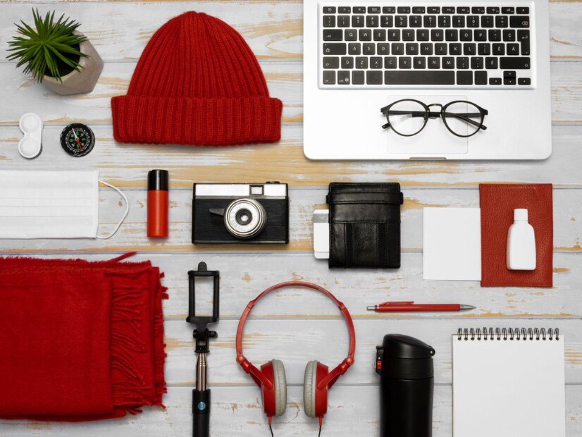 Assorted travel essentials including a laptop, camera, headphones, wallet, and personal items on a wooden surface.