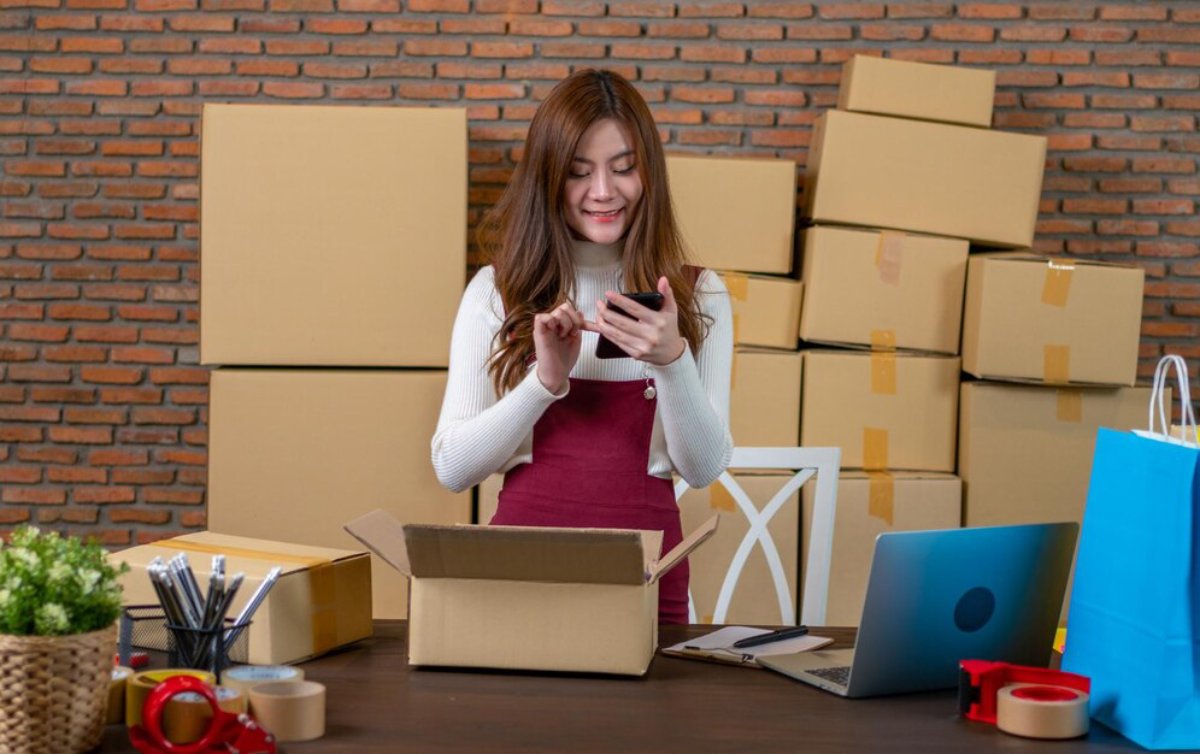 Woman in a red apron using a smartphone with many cardboard boxes and packing supplies around in a brick-walled room.