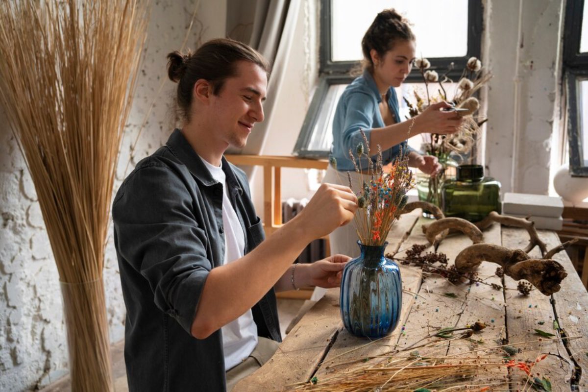 Two people arranging dried flowers and plants on a wooden table in a bright, artistic studio.