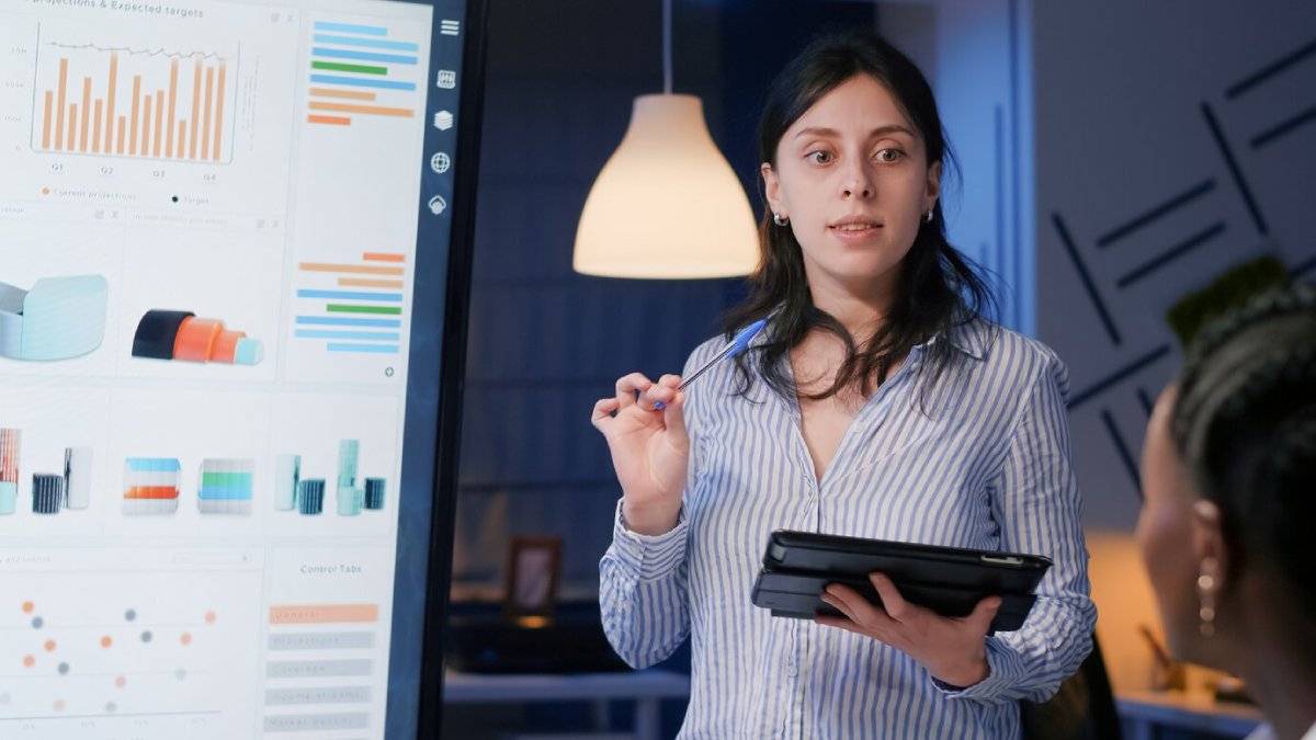 Woman presenting business data on digital screens to a colleague in an office setting.
