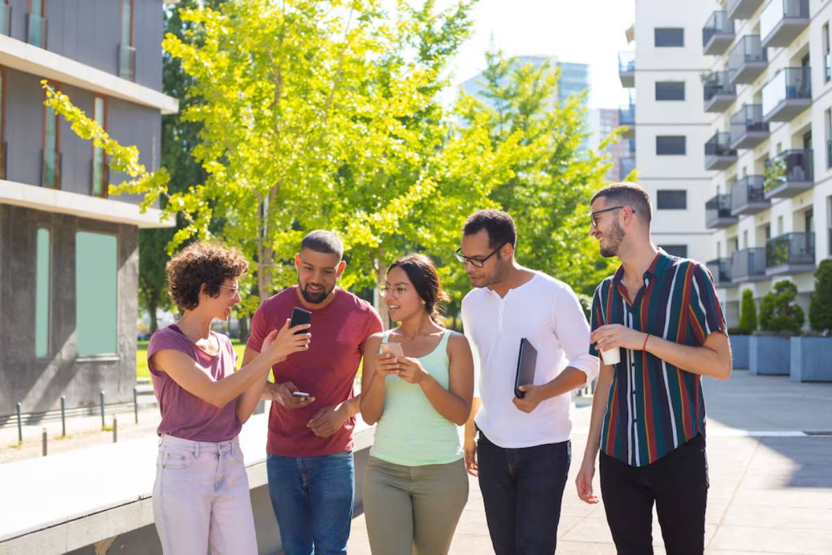 Group of five young adults chatting and walking outdoors in a sunny urban setting, with modern buildings and trees around.