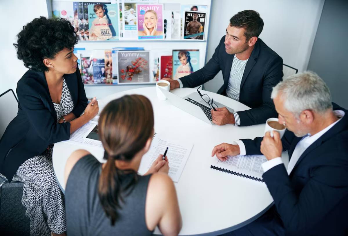 Four professionals sitting around a table in a modern office, discussing papers, with magazines on the wall behind them.