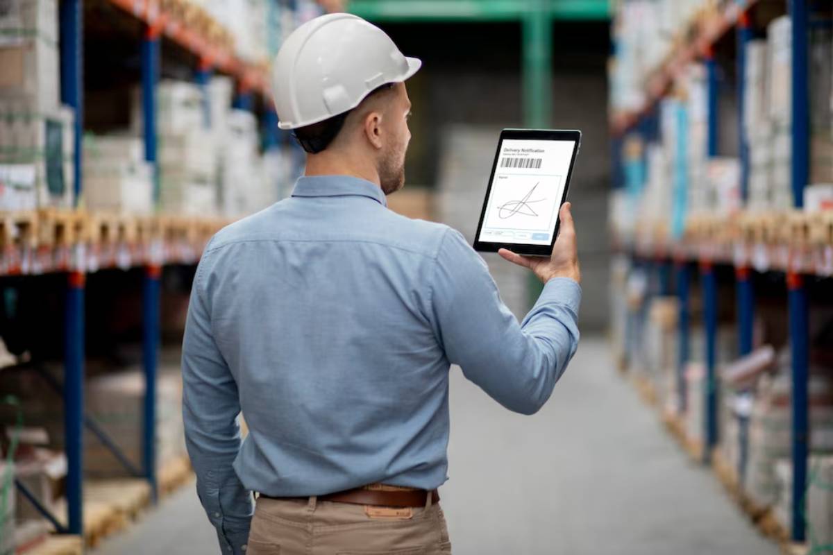 Man in hard hat using a tablet in a warehouse, reviewing a digital document.
