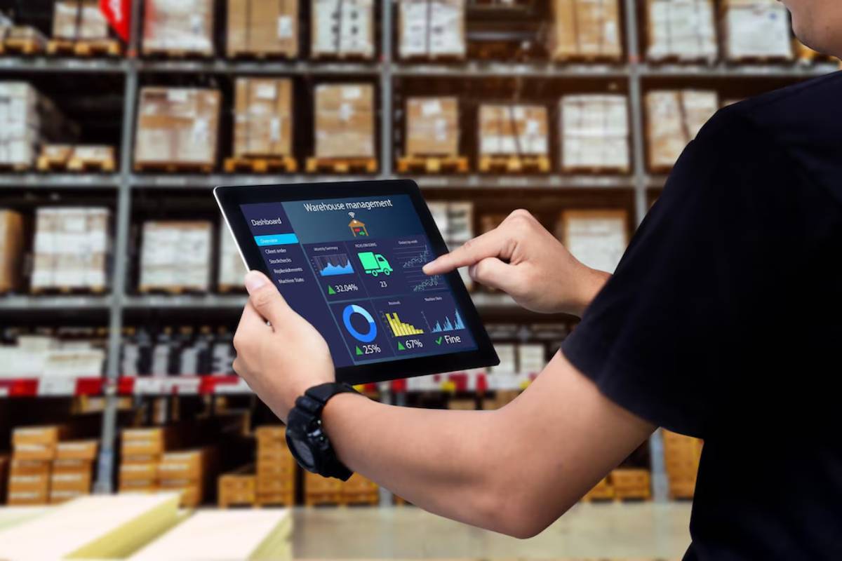 Man in warehouse uses a tablet displaying logistics data, with shelves of boxes in the background.