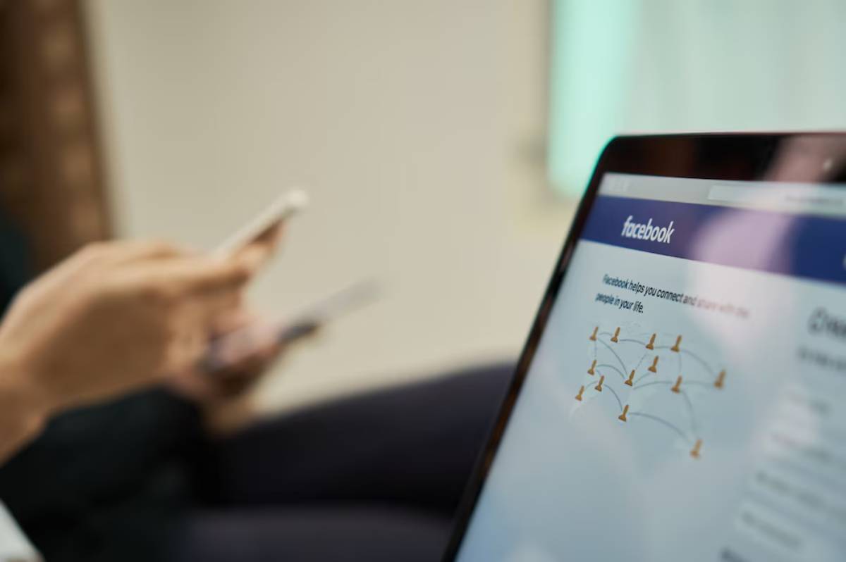 Person using smartphone with blurred Facebook page visible on tablet screen in foreground.