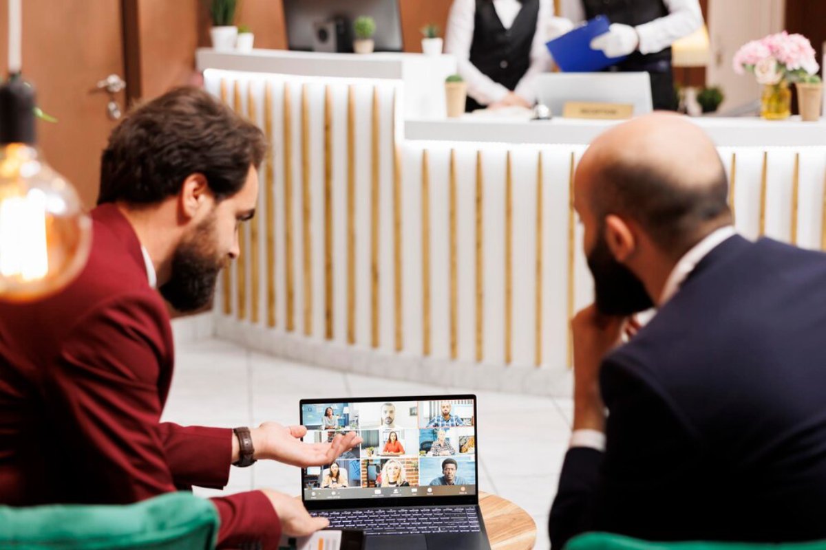 Two men in a modern office setting discussing over a laptop showing a virtual meeting with multiple participants.