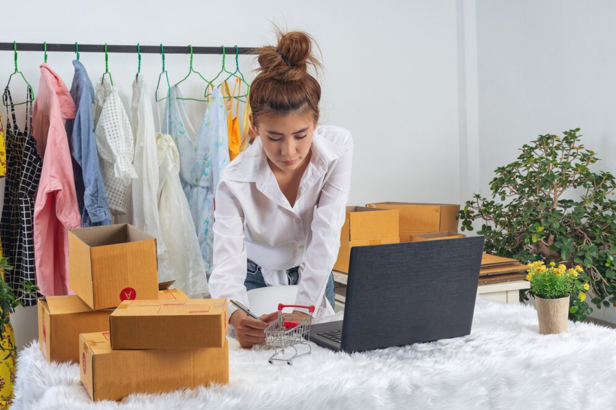 A woman organizing packages and working on a laptop in a home clothing business setup.