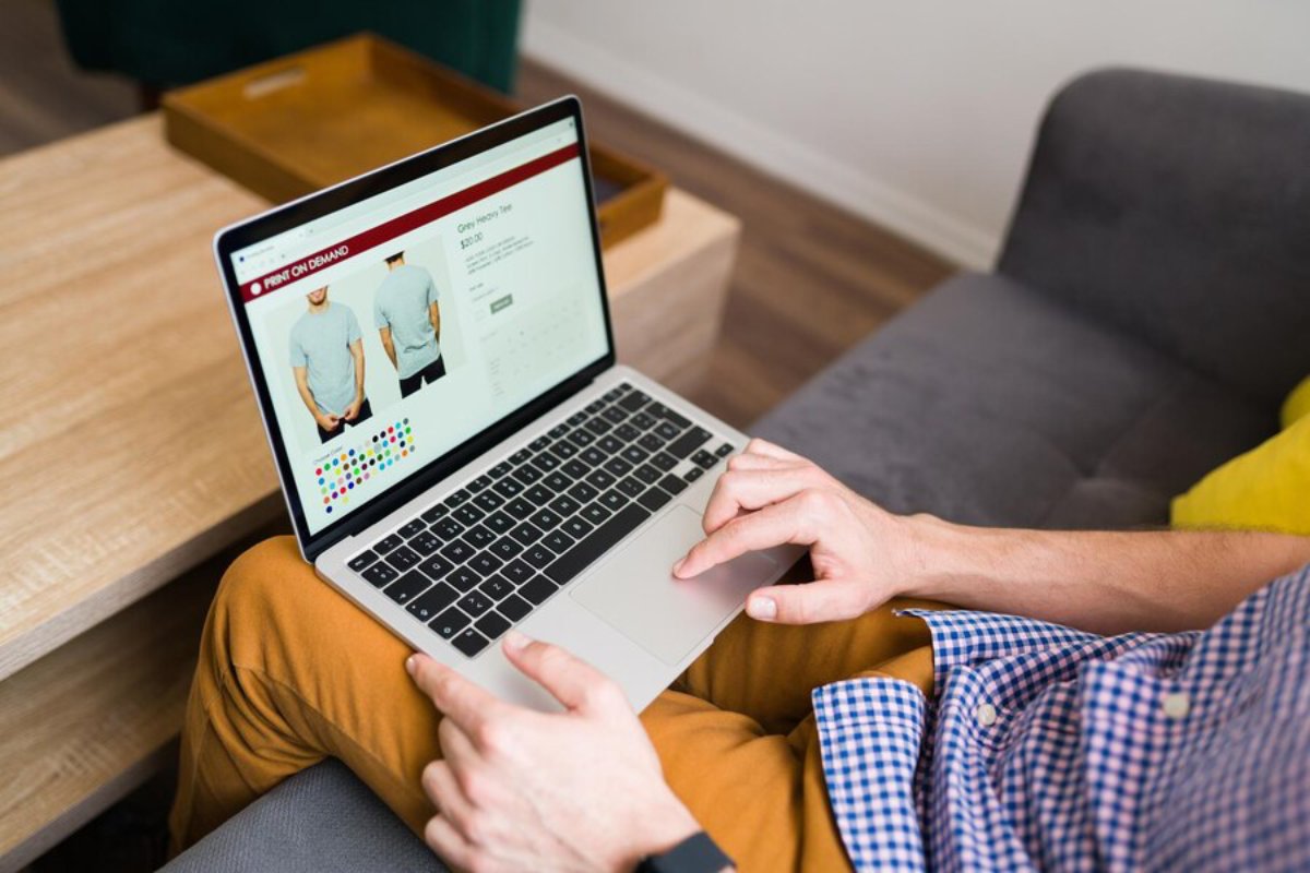 Man using a laptop to browse custom t-shirt printing options on a website while sitting on a sofa.