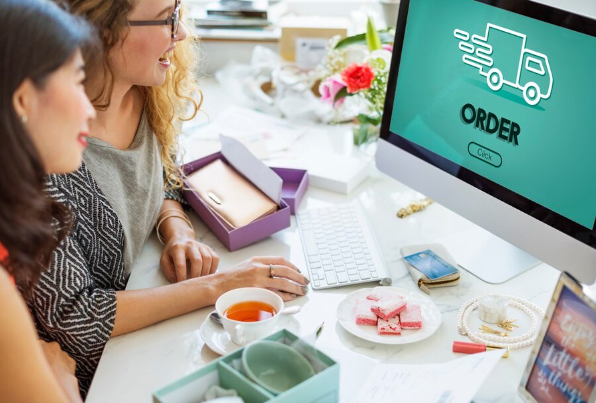 Two women review online delivery options on a computer screen in a cluttered office space filled with documents and items.