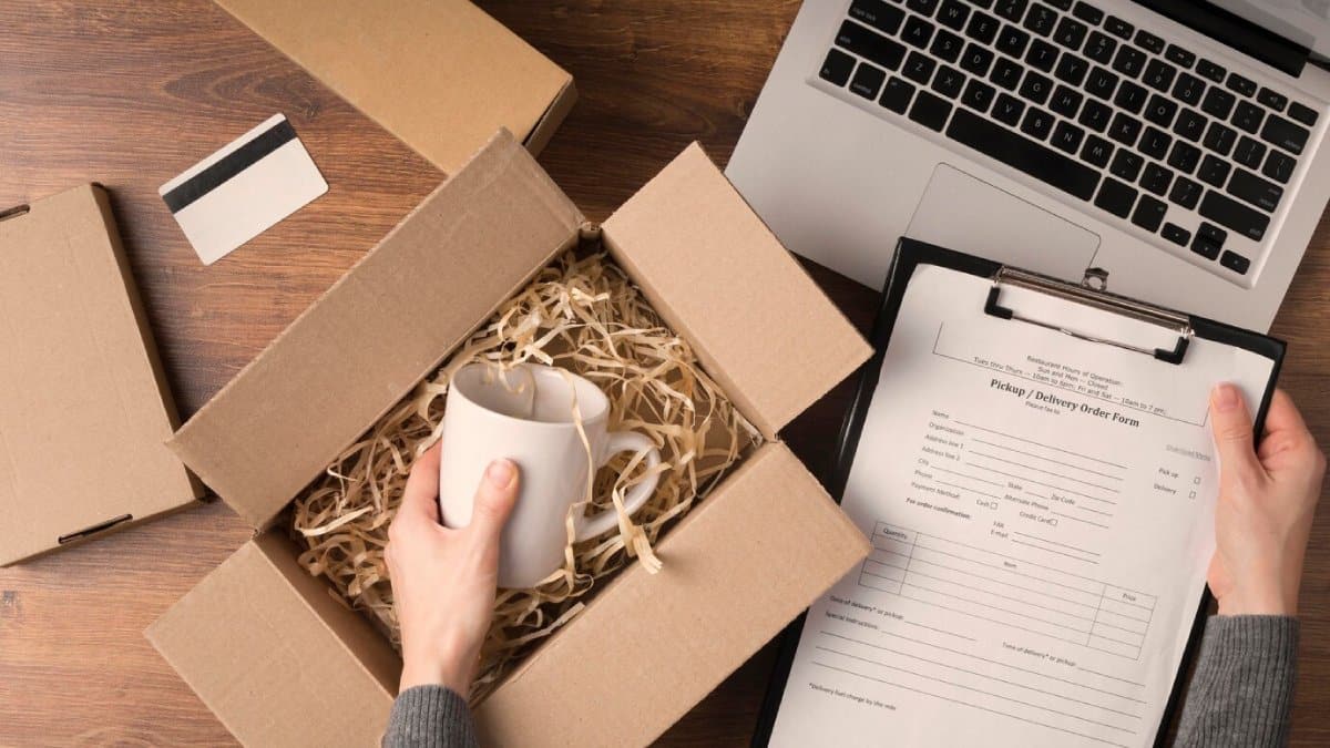Person unpacking a mug from a box, with a laptop and delivery form on a wooden table.