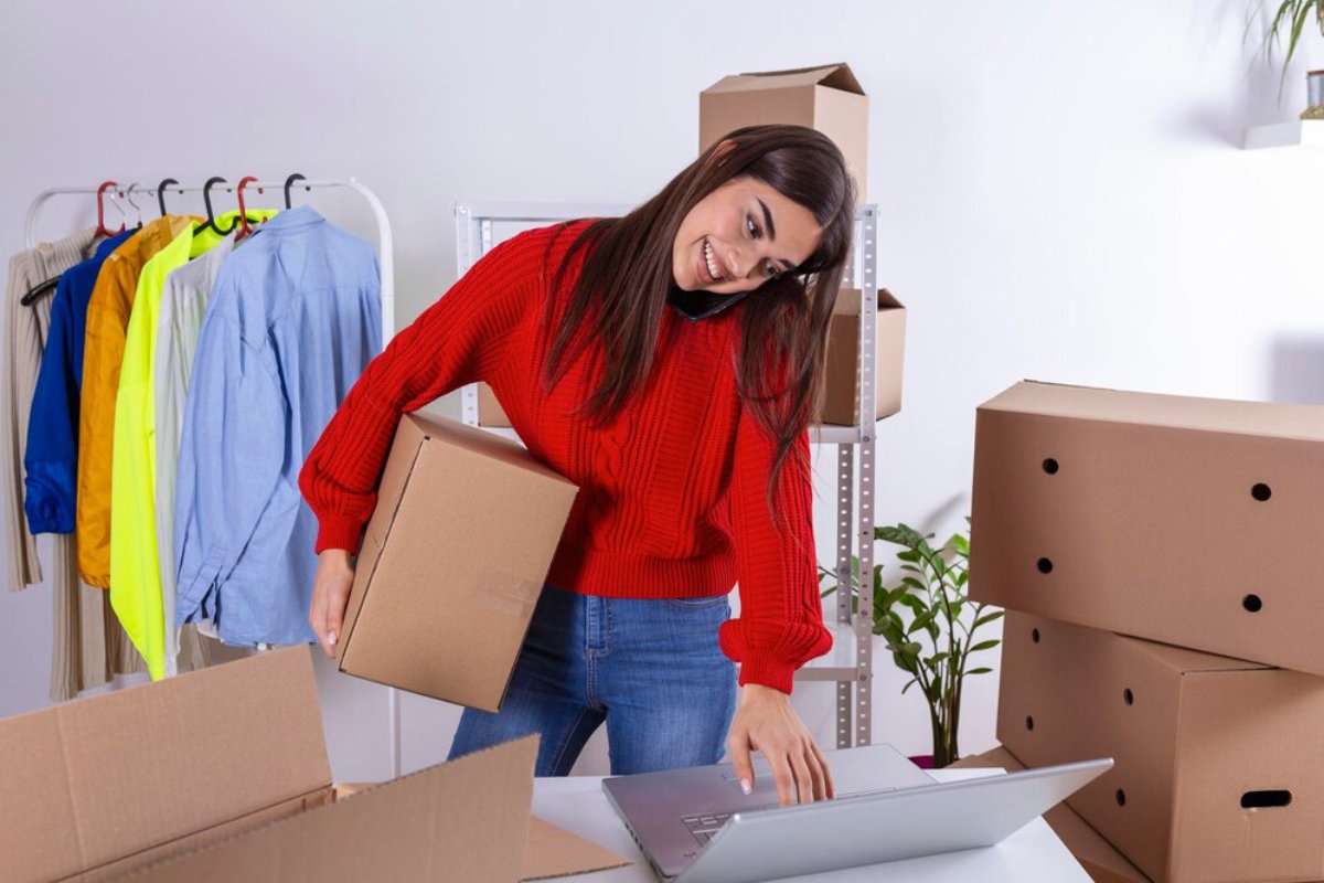 Woman in red sweater packing boxes and using laptop in a clothing warehouse.