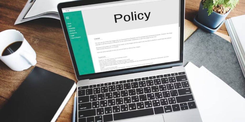 Laptop on wooden desk displaying a Policy document, with a coffee cup and plant beside it.