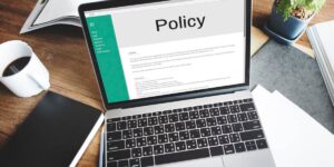 Laptop on wooden desk displaying a Policy document, with a coffee cup and plant beside it.