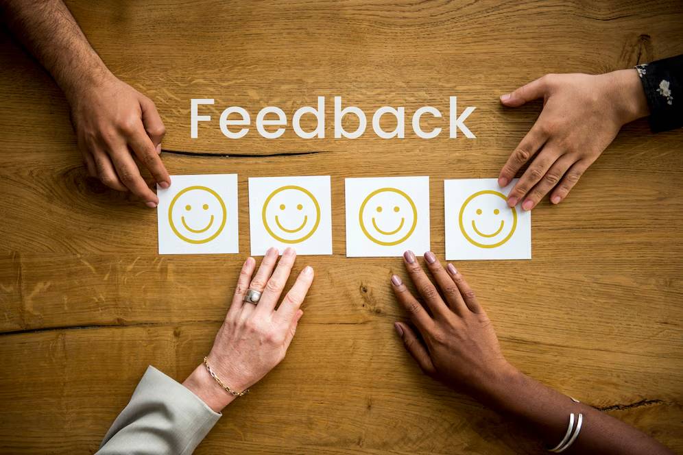 Four diverse hands pointing at smiley face cards on a wooden table labeled Feedback.
