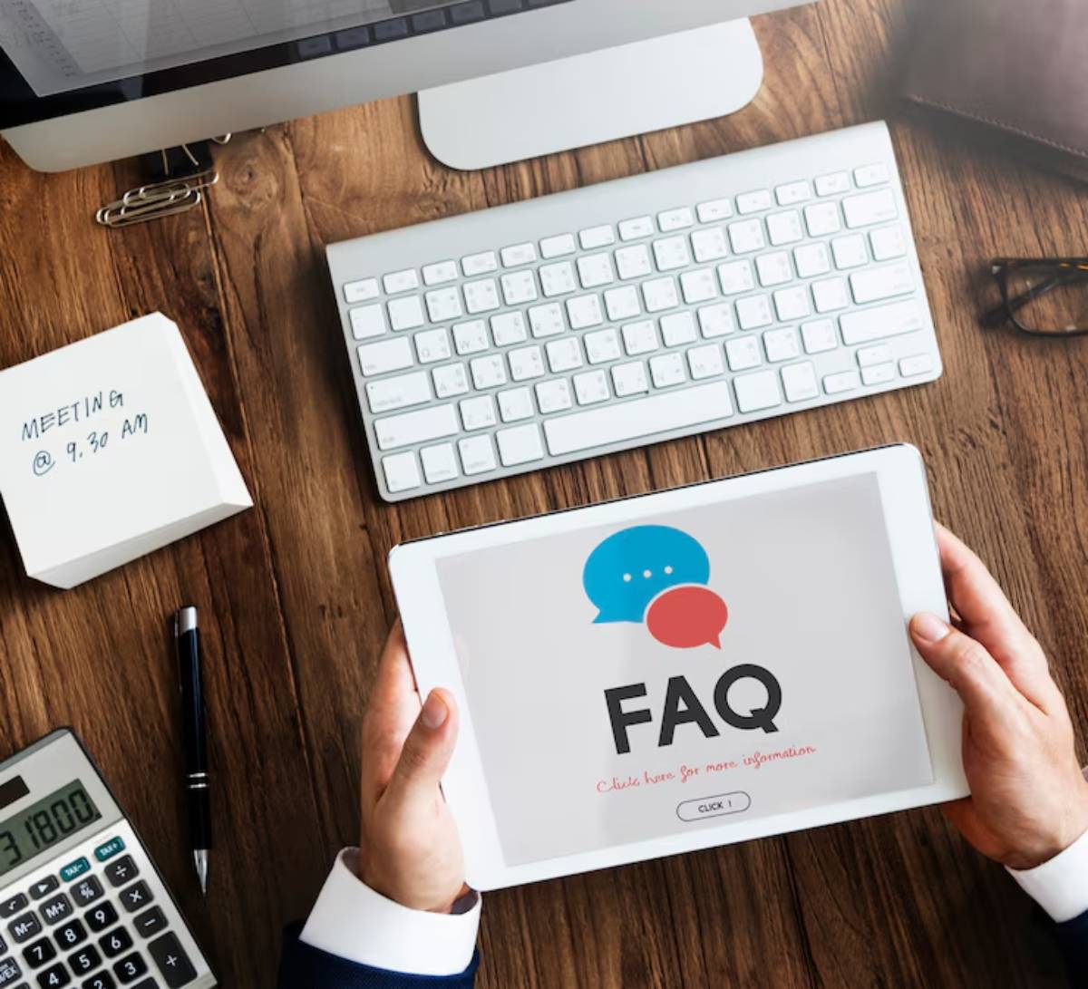 Person holding a tablet displaying FAQ with a desktop setup including computer, keyboard, and stationery on a wooden desk.