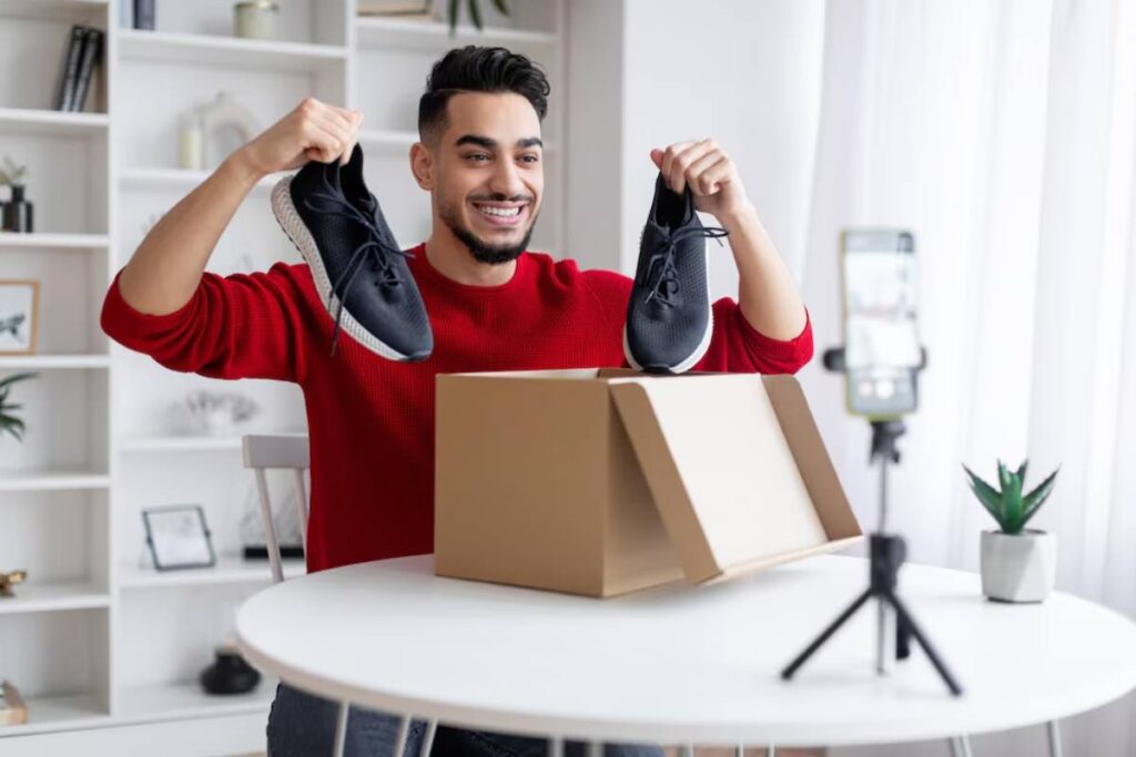 Man in red sweater smiling and holding up sneakers during a video blog session.