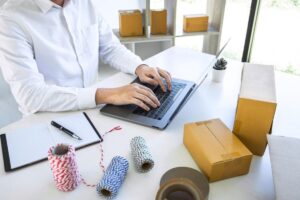Man in white shirt using laptop at desk with packing supplies and boxes for shipping.