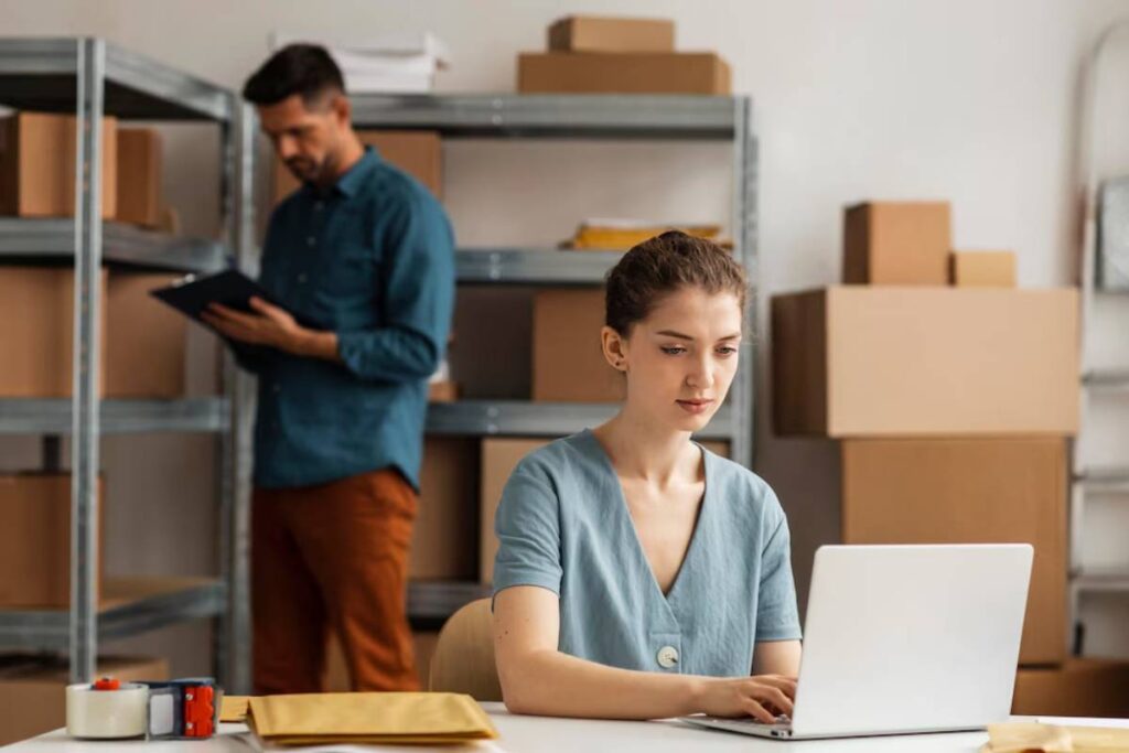 A woman works on a laptop in a storage room while a man reviews documents in the background.