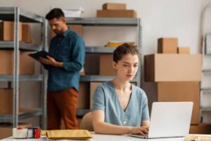 A woman works on a laptop in a storage room while a man reviews documents in the background.