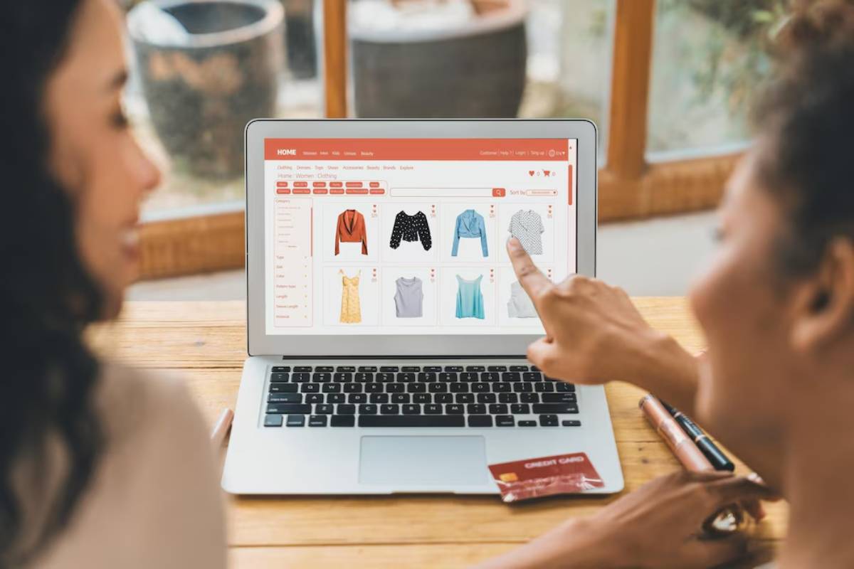 Two women shopping online on a laptop displaying clothing items, with a credit card in view.
