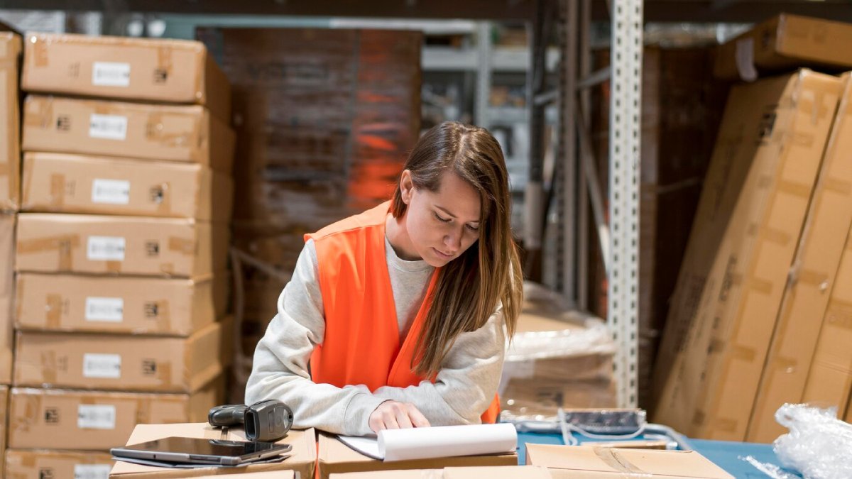 A woman in a safety vest reviews documents in a warehouse full of cardboard boxes.