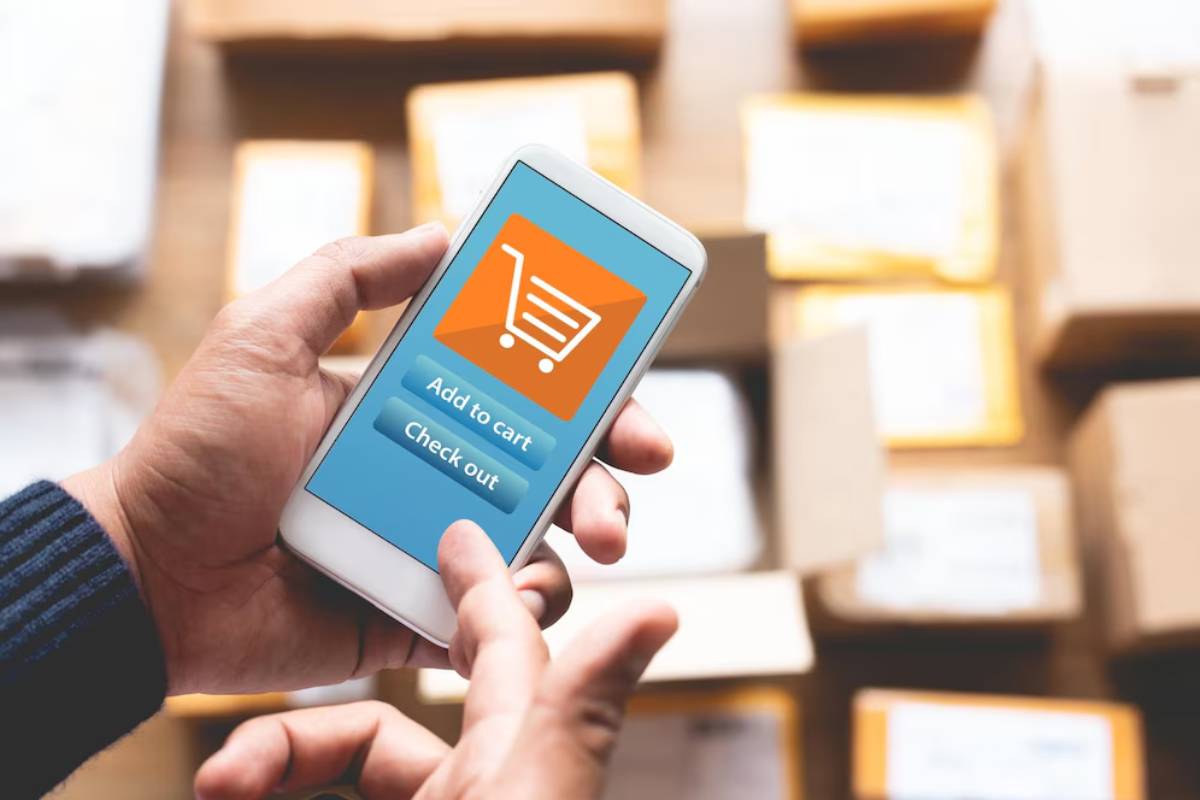 Hands holding a smartphone with an online shopping cart interface, surrounded by boxes in a warehouse.