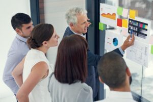 Five professionals discussing charts and data on a glass wall in a modern office.