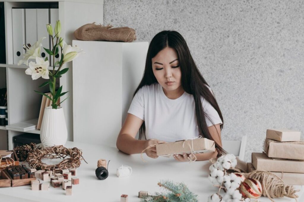 Woman wrapping gifts at a table decorated with craft supplies and seasonal decorations.