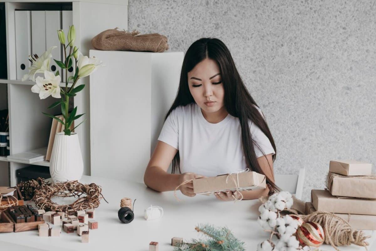 Woman wrapping gifts at a table decorated with craft supplies and seasonal decorations.