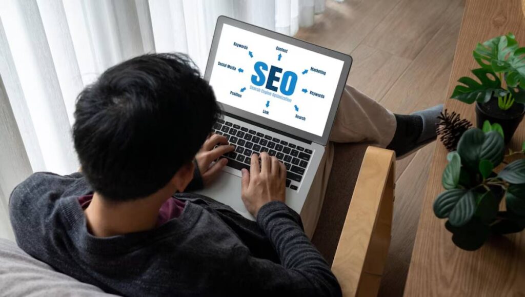 Man using a laptop displaying an SEO (Search Engine Optimization) diagram on the screen, sitting near a window with plants.