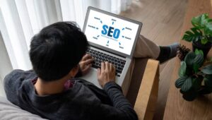 Man using a laptop displaying an SEO (Search Engine Optimization) diagram on the screen, sitting near a window with plants.