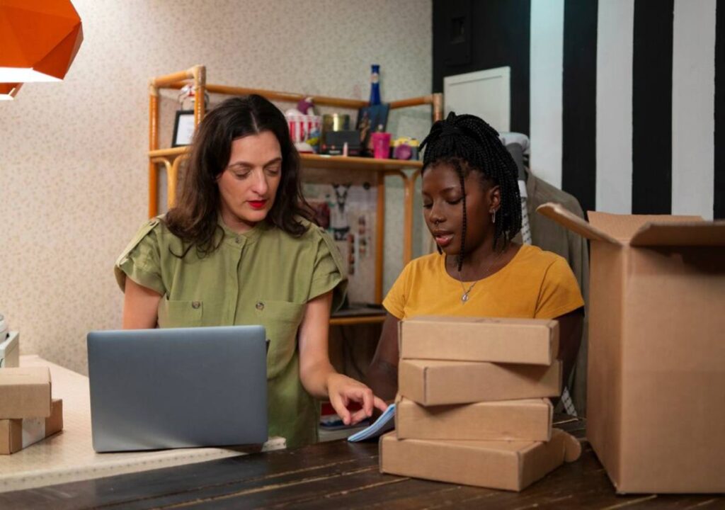 Two women working with a laptop and packages in a creatively cluttered office space.