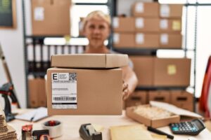Woman in a shipping warehouse with stacked boxes, focusing on a package in the foreground.