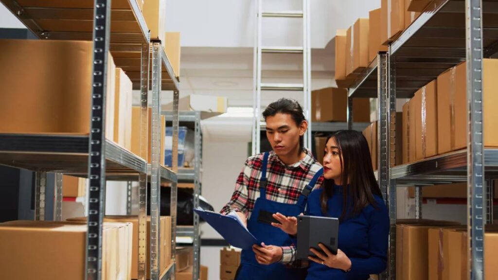 Two warehouse workers, a man and a woman, discussing inventory with a clipboard and tablet in a stockroom.