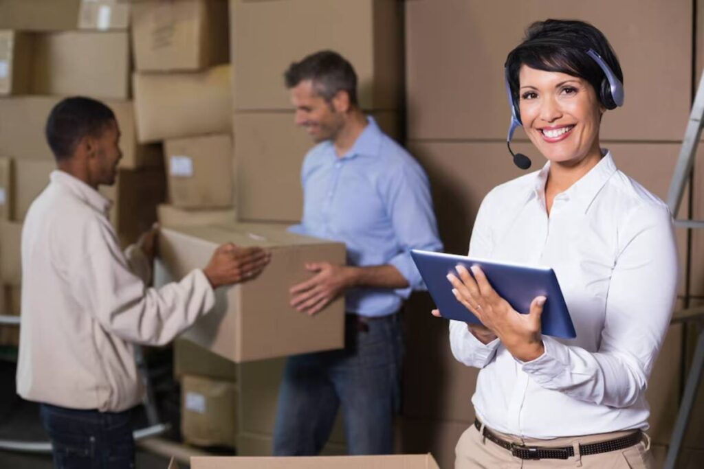 A woman with a headset and tablet smiles, overseeing two men handling boxes in a warehouse.