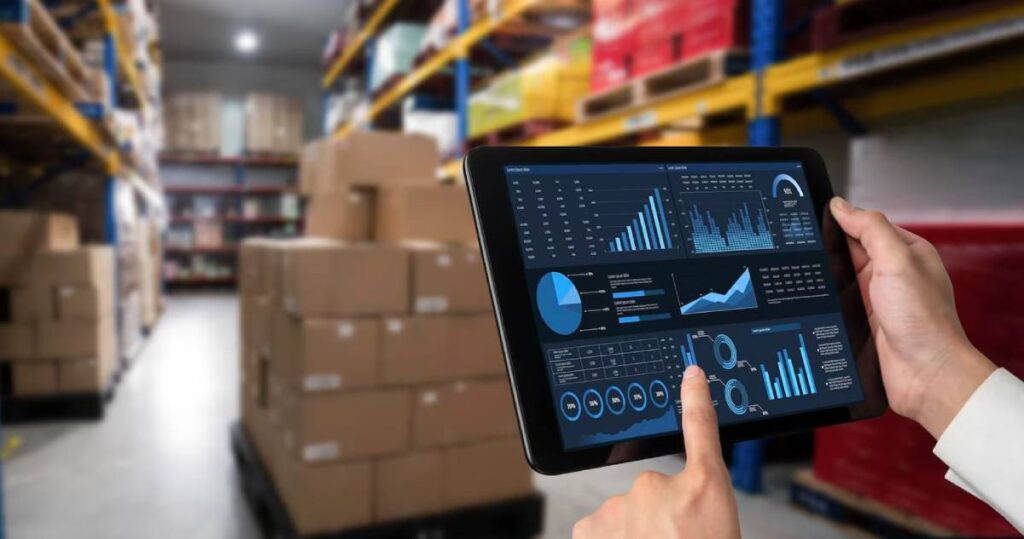 Hand holding a tablet displaying graphs and charts with a blurred warehouse background filled with boxes on shelves.
