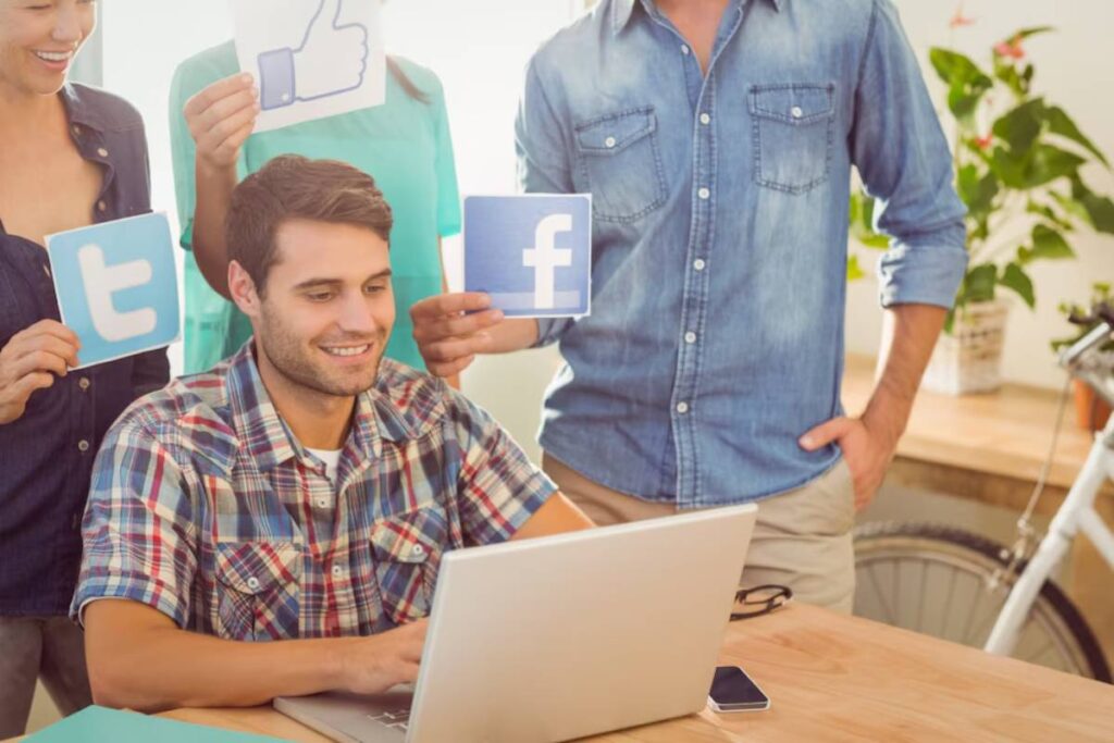 A young man uses a laptop while his colleagues hold up social media symbol cards like Facebook and Twitter around him.