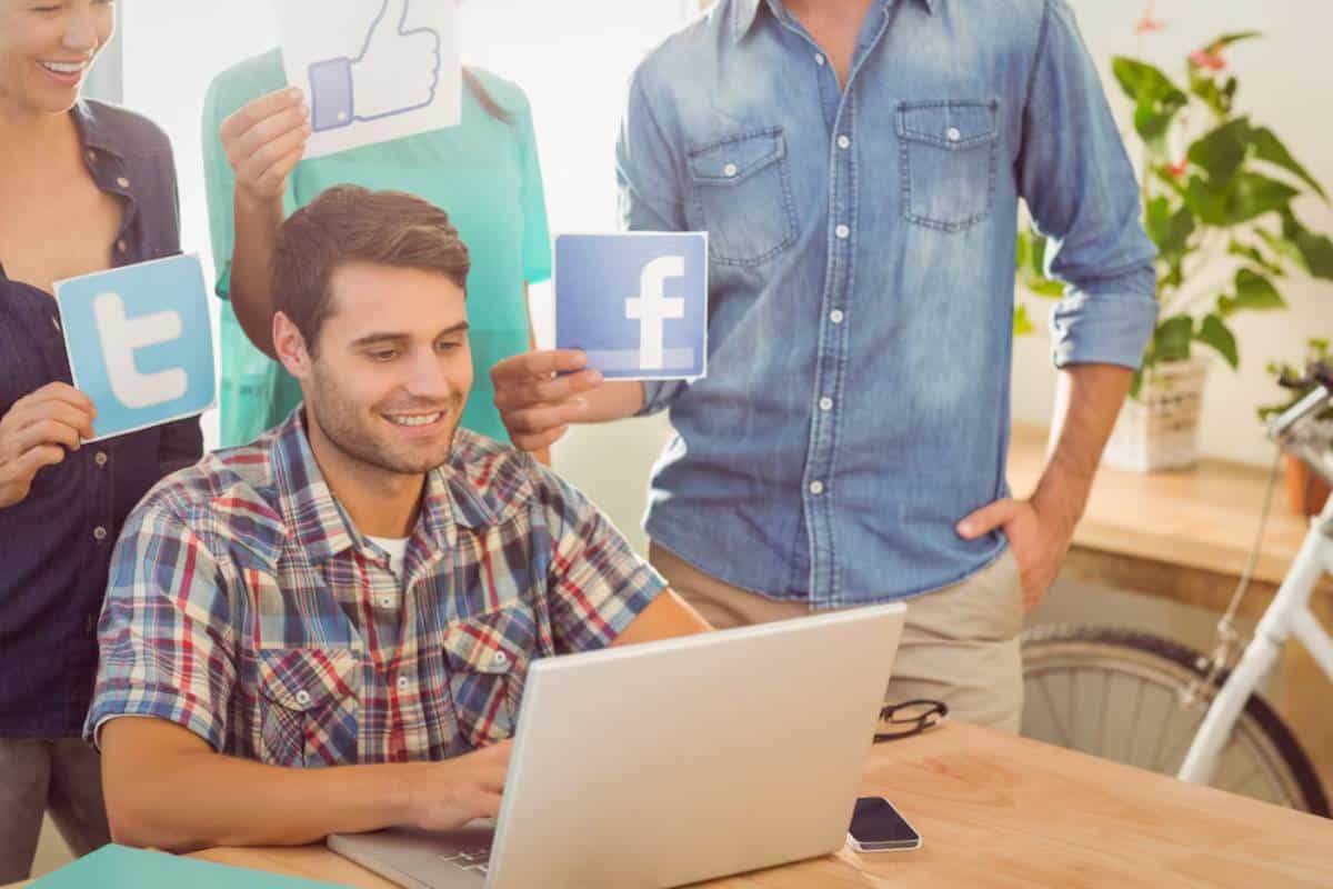 A young man uses a laptop while his colleagues hold up social media symbol cards like Facebook and Twitter around him.
