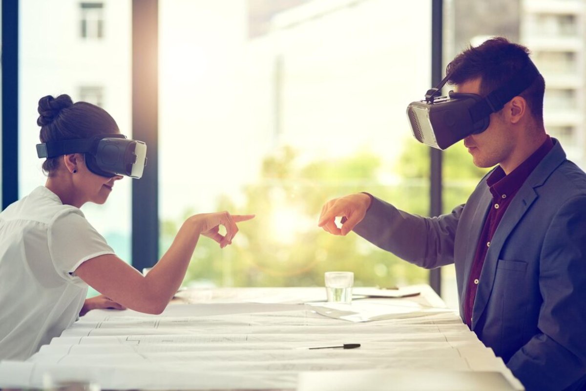 Two professionals wearing virtual reality headsets interacting over architectural plans in a sunlit office.