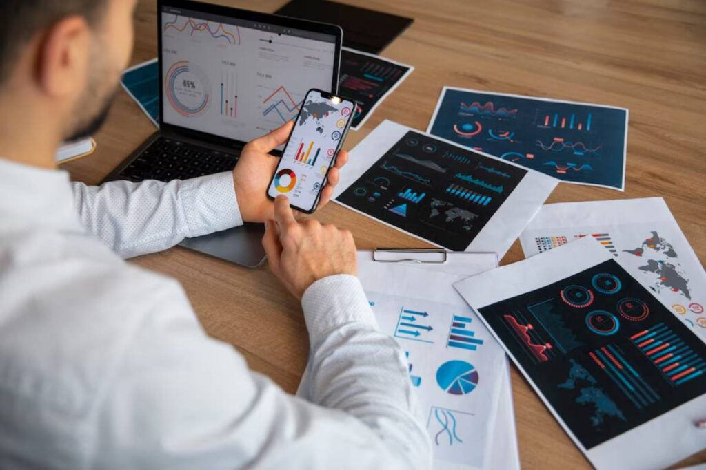 Businessman analyzing financial graphs on smartphone and laptop in an office setting.